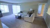 This single-floor space features a welcoming living room finished with wall-to-wall carpeting in a warm neutral tone that adds softness underfoot. The room is painted in a muted sage green, creating a calm and understated atmosphere throughout. A large sliding glass door fitted with vertical blinds serves as the focal point of the space, allowing an abundance of natural light to fill the room while providing direct access to an outdoor green area beyond.

The living area is generously sized, comfortably accommodating a full sofa and a separate armchair, both upholstered in light beige fabric and accented with decorative pillows. A dark-finished round coffee table with a lower shelf sits centrally between the seating pieces, offering both style and practical storage. A table lamp and small potted plant on a side table near the sofa contribute to the room's relaxed, livable feel.

White baseboards and trim provide clean contrast against the soft wall color, while a doorway visible on the right suggests additional rooms beyond the living area. Overall, the space conveys a practical, comfortable layout with ample natural light and a cohesive, neutral design palette.