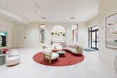 Located at 3440 SW 76 Terr in Davie, Florida, this beautifully designed single-floor amenity space offers a sophisticated and inviting environment across a generous footprint. The interior is defined by a crisp, all-white palette complemented by warm wood accents, large-format porcelain tile flooring, and abundant recessed lighting that creates a bright, airy ambiance throughout.

The central lobby and lounge area serves as the heart of the space, anchored by a bold circular crimson rug that grounds a curated seating arrangement of plush, curved sofas and lounge chairs. An elegant arched architectural feature adorned with lush moss wall art adds a distinctive design focal point. Floor-to-ceiling windows and glass entry doors allow generous natural light and provide views of the outdoor pool area.

A hallway corridor connects the various zones, including a fully equipped fitness center accessible via glass-paneled doors, a game room with a sleek billiard table framed by arched windows overlooking tropical greenery, and a semi-private media lounge defined by flowing white curtains and a wall-mounted television. A wet bar and kitchenette area features sage-green cabinetry, a large island with bar seating, and pendant lighting. Additional seating nooks and a curved red banquette complete this dynamic, resort-style amenity space.