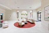 Located at 3440 SW 76 Terr in Davie, Florida, this beautifully designed single-floor amenity space offers a sophisticated and inviting environment across a generous footprint. The interior is defined by a crisp, all-white palette complemented by warm wood accents, large-format porcelain tile flooring, and abundant recessed lighting that creates a bright, airy ambiance throughout.

The central lobby and lounge area serves as the heart of the space, anchored by a bold circular crimson rug that grounds a curated seating arrangement of plush, curved sofas and lounge chairs. An elegant arched architectural feature adorned with lush moss wall art adds a distinctive design focal point. Floor-to-ceiling windows and glass entry doors allow generous natural light and provide views of the outdoor pool area.

A hallway corridor connects the various zones, including a fully equipped fitness center accessible via glass-paneled doors, a game room with a sleek billiard table framed by arched windows overlooking tropical greenery, and a semi-private media lounge defined by flowing white curtains and a wall-mounted television. A wet bar and kitchenette area features sage-green cabinetry, a large island with bar seating, and pendant lighting. Additional seating nooks and a curved red banquette complete this dynamic, resort-style amenity space.