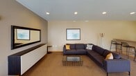 This single-floor space in West St. Paul, Minnesota, is thoughtfully designed to serve multiple functions within one open layout. The floor plan flows naturally from a functional kitchen into a shared living and meeting area, creating a versatile environment suited for both casual gatherings and structured activities.

The kitchen is outfitted with warm honey-toned wood cabinetry, a double-basin sink, full-size refrigerator, electric range, microwave, and ample counter space. Tile flooring and generous storage make it a practical, fully equipped cooking area. A breakfast bar separates the kitchen from the adjoining space, providing additional seating and a visual transition between zones.

The lounge area features a large sectional sofa in deep charcoal, accented with mustard and patterned throw pillows, anchored by a wire-frame coffee table over a woven area rug. A wall-mounted flat-screen television sits above a sleek white floating credenza, complemented by framed landscape artwork and recessed lighting overhead.

A dedicated meeting or dining zone accommodates folding tables and chairs, positioned near large windows fitted with vertical blinds that allow generous natural light to fill the space. The overall ambiance is clean and neutral, with light beige walls, carpet, and modern furnishings that create a welcoming, functional shared environment.