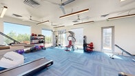 Located in Colorado Springs, CO, this fitness center offers a thoughtfully designed workout space that balances functionality with a clean, modern aesthetic. The single open-plan room is finished with distinctive blue herringbone-patterned carpet flooring and crisp white walls, creating a bright and energizing environment. Multiple large windows line the perimeter, flooding the space with abundant natural light and providing views of the surrounding Colorado landscape, including distant mountain vistas. Overhead, wood-accented linear pendant lights and multiple ceiling fans ensure the space remains well-lit and comfortable during workouts. The facility is comprehensively equipped to support a wide range of fitness activities. Cardio options include treadmills, a stair climber, and a stationary bike, while strength training needs are addressed through multi-station cable machines, a full dumbbell rack, and various benches. A dedicated functional training area features medicine balls, resistance bands, yoga mats, and stability balls neatly organized on freestanding racks. Full-length mirrors along one wall enhance the sense of space and allow users to monitor their form. Direct access doors open to an outdoor area, further connecting the fitness center to the surrounding environment. The overall ambiance is clean, airy, and well-maintained, making it a practical and inviting space for daily exercise.
