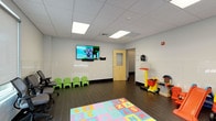 This single-floor space is designed as a dedicated children's play and waiting area, combining functionality with a child-friendly environment. The room features dark tile flooring throughout, providing a durable and easy-to-maintain surface, while a drop ceiling with recessed panel lighting ensures consistent, bright illumination across the entire space.

The play area is generously equipped with a variety of age-appropriate toys and activities, including a ride-on car, a miniature slide, a small basketball hoop, a playhouse, and a toy kitchen set, offering children multiple ways to stay engaged. A colorful foam alphabet and number puzzle mat defines the central play zone, adding both visual interest and a soft surface for younger children.

A wall-mounted flat-screen television plays children's programming, providing additional entertainment. Small green chairs line one wall, offering seating scaled for young children, while adult-sized office chairs positioned near windows accommodate accompanying caregivers. The windows, fitted with roller shades, allow natural light to filter into the space. The neutral white walls and clean, modern aesthetic contribute to an organized and welcoming atmosphere suited for families.