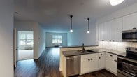 Located at Greybarn Amityville on Broadway in Amityville, NY, this single-floor residence offers a thoughtfully designed open-concept layout that seamlessly connects the kitchen and living areas. The kitchen is a standout feature, showcasing white shaker-style cabinetry with dark hardware, dark countertops, a classic white subway tile backsplash, and a full suite of stainless steel appliances including a refrigerator, range, over-the-range microwave, and dishwasher. A peninsula with pendant lighting serves as both a workspace and a visual anchor between the kitchen and the expansive living area. Rich, dark hardwood-style flooring flows throughout the main living spaces, adding warmth and contrast to the bright white walls. The generously sized living area is bathed in natural light from large windows. The bedroom features soft carpet flooring, a wide closet, and a large window with outdoor views. An in-unit stacked washer and dryer adds everyday convenience. The bathroom is finished with floor-to-ceiling white subway tile, a tub-shower combination, a wide vanity with dark countertops, white cabinetry, and a large mirror, creating a clean and polished aesthetic throughout.