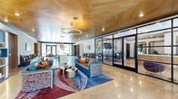 Located at 850 Ronald Reagan Drive in Evans, Georgia, this single-story clubhouse offers a generously sized, thoughtfully designed community gathering space. The layout encompasses two living areas and a connecting hallway, all unified by a bold, contemporary design aesthetic.

The main living area commands immediate attention with its striking herringbone wood-paneled ceiling, polished concrete floors, and an elongated marble-topped bar island flanked by industrial-style barstools. A cascading glass pendant chandelier serves as a dramatic focal point, while built-in banquette seating with vibrant teal, orange, and gold accent pillows lines one wall beneath a wall-mounted display. Eclectic, patterned lounge chairs and colorful area rugs complete the lively, curated atmosphere. Large glass-paneled doors and expansive windows flood the space with natural light.

The second living area offers a more intimate ambiance, featuring warm wood flooring, a teal-painted ceiling, sputnik-style brass chandeliers, and a glossy teal tile wet bar. Plush, curved neutral sofas and blue accent chairs surround a rustic wood coffee table, creating an inviting lounge environment. Windows along the perimeter provide views of the surrounding exterior. Together, both areas deliver a vibrant, upscale clubhouse experience well-suited for community gatherings and social events.