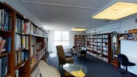 This single-floor library and reading space is designed to provide a welcoming, resource-rich environment for reading and study. The room is lined with tall, dark-wood bookshelves filled with an extensive and diverse collection of books, spanning multiple rows and organized neatly across the walls and in freestanding shelf units throughout the space. The flooring is covered in a dark, textured carpet that adds warmth and helps define the reading areas. Natural light filters in through large windows along one wall, offering views of the surrounding urban environment and brightening the interior. Overhead, flush-mounted panel lighting supplements the natural light, ensuring the space remains well-lit throughout. Comfortable seating is thoughtfully arranged throughout, including cushioned armchairs and a sofa grouped around a glass-topped coffee table, creating intimate nooks ideal for leisurely reading. Additional seating, including side chairs and small tables, is positioned near the windows to take advantage of the natural light. The overall ambiance is quiet and studious, with a functional yet comfortable layout that balances ample storage for materials with inviting spaces for patrons to sit, browse, and engage with the collection.
