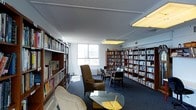 This single-floor library and reading space is designed to provide a welcoming, resource-rich environment for reading and study. The room is lined with tall, dark-wood bookshelves filled with an extensive and diverse collection of books, spanning multiple rows and organized neatly across the walls and in freestanding shelf units throughout the space. The flooring is covered in a dark, textured carpet that adds warmth and helps define the reading areas. Natural light filters in through large windows along one wall, offering views of the surrounding urban environment and brightening the interior. Overhead, flush-mounted panel lighting supplements the natural light, ensuring the space remains well-lit throughout. Comfortable seating is thoughtfully arranged throughout, including cushioned armchairs and a sofa grouped around a glass-topped coffee table, creating intimate nooks ideal for leisurely reading. Additional seating, including side chairs and small tables, is positioned near the windows to take advantage of the natural light. The overall ambiance is quiet and studious, with a functional yet comfortable layout that balances ample storage for materials with inviting spaces for patrons to sit, browse, and engage with the collection.