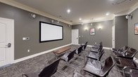 This dedicated screening room is designed to deliver an immersive viewing experience in a refined, intimate setting. The space is finished with smooth grey walls accented by crisp white crown molding and baseboards, lending a polished, cinematic atmosphere throughout. A large, black-framed projection screen is prominently mounted on the front wall, providing a commanding focal point for the room. A ceiling-mounted projector is positioned toward the rear, ensuring clear, high-quality projection across the full screen surface.

Seating consists of multiple dark leather chairs with chrome frames arranged in rows, offering comfortable, stylish accommodation for a group of viewers. A tufted brown leather bench at the center of the room serves as an additional seating or footrest option. The floor is covered in a durable, patterned tile-style carpet that adds texture and visual interest while contributing to the room's acoustic qualities.

Wall-mounted sconce lighting provides warm, ambient illumination, and recessed ceiling lights supplement the overall brightness. Multiple surround sound speakers are mounted at various points along the walls, ensuring balanced audio throughout the space. A curated gallery of framed vintage automotive and aviation posters adds a distinctive thematic character to the room, completing its well-appointed, purposeful design.