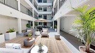 Located at 3050 W 11th St in Los Angeles, California, this inviting space centers around a beautifully designed open-air courtyard that serves as a shared living area. The multi-story building surrounds the courtyard on all sides, with upper-level balconies and railings visible above, creating a dramatic atrium-style atmosphere that draws in abundant natural light from above. String lights are strung overhead, adding a warm, ambient glow for evening gatherings.

The courtyard living area is thoughtfully furnished with modern, neutral-toned lounge chairs, ottomans, and a dining set complete with a shaded umbrella table — ideal for both relaxed lounging and casual dining. Woven area rugs anchor the seating zones, adding texture and definition to the open floor space. Lush tropical plants, including tall palms, fiddle-leaf figs, and curated planter arrangements, are strategically placed throughout, bringing vibrant greenery and a resort-like feel to the space.

The surrounding walls are finished in crisp white stucco, complemented by clean grey doors and large windows that maintain a cohesive, contemporary aesthetic. The smooth concrete flooring ties the entire space together with a minimalist, modern sensibility. Overall, the courtyard exudes a bright, airy, and welcoming California ambiance well-suited for community living.