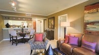 This single-floor residence showcases an open-concept layout that seamlessly connects the living, dining, and kitchen areas, creating a cohesive and functional living environment. The walls are finished in warm golden-tan tones, complemented by crisp white crown molding that adds a refined architectural detail throughout the space.

The living area is anchored by a rich brown leather sofa accented with vibrant pink and purple throw pillows, paired with a patterned accent chair featuring bold red cushions. A tall floor lamp provides warm ambient lighting, enhancing the inviting atmosphere. Light-toned tile flooring extends throughout the main living spaces, offering both durability and a clean aesthetic.

Adjacent to the living area, the dining space accommodates a dark wood table surrounded by upholstered chairs, ideal for everyday dining or entertaining. A framed artwork piece and a dark wood sideboard add character and storage to the space.

The kitchen, visible in the background, features light cabinetry and appears well-appointed. An interior door leads to additional rooms, suggesting a practical and well-organized floor plan. Overall, the space presents a warm, elegantly styled environment with a cohesive color palette.