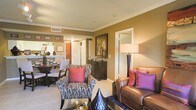 This single-floor residence showcases an open-concept layout that seamlessly connects the living, dining, and kitchen areas, creating a cohesive and functional living environment. The walls are finished in warm golden-tan tones, complemented by crisp white crown molding that adds a refined architectural detail throughout the space.

The living area is anchored by a rich brown leather sofa accented with vibrant pink and purple throw pillows, paired with a patterned accent chair featuring bold red cushions. A tall floor lamp provides warm ambient lighting, enhancing the inviting atmosphere. Light-toned tile flooring extends throughout the main living spaces, offering both durability and a clean aesthetic.

Adjacent to the living area, the dining space accommodates a dark wood table surrounded by upholstered chairs, ideal for everyday dining or entertaining. A framed artwork piece and a dark wood sideboard add character and storage to the space.

The kitchen, visible in the background, features light cabinetry and appears well-appointed. An interior door leads to additional rooms, suggesting a practical and well-organized floor plan. Overall, the space presents a warm, elegantly styled environment with a cohesive color palette.