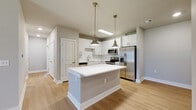 Located at 3795 Bismarck Ct in Oxford, Florida, this single-story residence presents a clean, contemporary design throughout its well-appointed interior. The kitchen serves as a central highlight, showcasing crisp white shaker-style cabinetry extending to the ceiling, a classic white subway tile backsplash, and a full suite of stainless steel appliances including a French door refrigerator, electric range, built-in microwave, and dishwasher. A generously sized center island with a bright white countertop anchors the space, illuminated by a pair of brushed brass pendant lights that add a warm, refined accent. Light wood-tone flooring runs throughout the kitchen and connecting areas, enhancing the airy, modern aesthetic. The bedrooms feature neutral-toned carpet, ceiling fans for comfort, and windows that allow natural light to filter in. Closet access is conveniently integrated into the bedroom layout. The bathroom is finished with a wide-format vanity featuring a white countertop and brushed nickel hardware, a large mirror, and a soaking tub enclosed by floor-to-ceiling white subway tile. Neutral gray walls and clean white trim are consistent throughout, contributing to a cohesive, move-in-ready atmosphere.