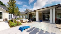 Located at 115 Dean Ave in Franklin, Massachusetts, this single-story amenity space is thoughtfully designed to blend indoor comfort with outdoor recreation. The exterior presents a craftsman-inspired aesthetic, combining sage-toned lap siding, stone accents, and a pitched shingle roof. Covered outdoor areas extend from the main structure, supported by clean white columns and exposed wood beam details that carry through to the interior.

Inside, the lounge area features soaring vaulted ceilings with exposed wooden beams and a floor-to-ceiling tiled fireplace flanked by a wall-mounted television. An orb chandelier and vibrant artwork contribute to the contemporary, inviting atmosphere. The space is furnished with stylish seating arrangements centered around a concrete coffee table, all anchored by layered area rugs.

The covered patio transitions seamlessly to the outdoors and includes a dining area with ample seating, perfect for gatherings. An outdoor kitchen with stone surround and a pergola overhead adds functional charm to the exterior. A paved courtyard with cornhole boards encourages casual outdoor activity.

Adjacent to the main building, a swimming pool is enclosed by black metal fencing and surrounded by lounge chairs, with lush landscaping providing a natural, park-like buffer throughout the grounds.