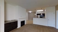 This single-floor residence showcases an open-concept layout that connects the living, dining, and kitchen areas into one cohesive space. The living area is anchored by a classic white-mantled fireplace with a sleek electric insert, flanked by dark cabinetry with granite detailing, adding a refined contrast to the neutral-toned walls and plush carpeted flooring.

The kitchen is positioned along one side of the space and features dark cabinetry, granite countertops, and stainless steel appliances. A raised breakfast bar peninsula separates the kitchen from the adjoining living and dining areas, offering both casual seating and additional workspace. Pendant lighting hangs above the bar, complementing the flush-mount ceiling fixtures that provide consistent, even illumination throughout.

The transition from carpeted living space to hardwood-style flooring in the kitchen and adjoining areas creates a natural visual boundary between zones. The neutral color palette of whites and warm grays throughout the space lends a clean, modern aesthetic, while the open layout ensures a sense of spaciousness and easy movement between areas.