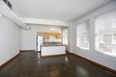 This single-floor unit showcases a modern loft-inspired design characterized by polished concrete floors, exposed ductwork, and crisp white walls throughout the main living areas. The open-plan layout connects the living space seamlessly to the kitchen, creating a generous, airy atmosphere well-suited for both relaxing and entertaining.

The kitchen is a functional centerpiece, featuring a peninsula island with a built-in cooktop and a grey laminate countertop, providing ample workspace and a natural gathering point. Warm-toned maple cabinetry lines the walls, complemented by stainless steel appliances including a full-size refrigerator. A dedicated sink area and additional counter space round out the kitchen's practical offerings. Large windows flood the kitchen and living area with natural light, offering views of the surrounding urban brick architecture.

The bedroom offers a change of texture with soft carpeted flooring and a ceiling fan fixture, providing comfort and warmth. A window frames city views, enhancing the room's connection to the urban environment.

The bathroom is well-appointed with a full tub and shower combination, a wide vanity featuring warm wood cabinetry, a large mirror with overhead lighting, and polished concrete flooring that ties back to the overall aesthetic. An adjacent doorway provides access to a walk-in closet, adding practical storage to complete this urban dwelling.