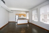 This single-floor unit showcases a modern loft-inspired design characterized by polished concrete floors, exposed ductwork, and crisp white walls throughout the main living areas. The open-plan layout connects the living space seamlessly to the kitchen, creating a generous, airy atmosphere well-suited for both relaxing and entertaining.

The kitchen is a functional centerpiece, featuring a peninsula island with a built-in cooktop and a grey laminate countertop, providing ample workspace and a natural gathering point. Warm-toned maple cabinetry lines the walls, complemented by stainless steel appliances including a full-size refrigerator. A dedicated sink area and additional counter space round out the kitchen's practical offerings. Large windows flood the kitchen and living area with natural light, offering views of the surrounding urban brick architecture.

The bedroom offers a change of texture with soft carpeted flooring and a ceiling fan fixture, providing comfort and warmth. A window frames city views, enhancing the room's connection to the urban environment.

The bathroom is well-appointed with a full tub and shower combination, a wide vanity featuring warm wood cabinetry, a large mirror with overhead lighting, and polished concrete flooring that ties back to the overall aesthetic. An adjacent doorway provides access to a walk-in closet, adding practical storage to complete this urban dwelling.