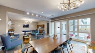 Located at 1010 S Pearl Expy in Dallas, TX, this single-floor community amenity space offers a thoughtfully designed, open-concept layout that blends entertainment and comfort seamlessly. The polished concrete floors and neutral warm-toned walls create a clean, modern backdrop throughout the entire space.

At the heart of the room sits a full-size billiards table beneath a striking mid-century modern chandelier with brass and globe accents. A wall-mounted flat-screen TV and wall sconces complete the game area, which is further supported by a wet bar and cabinetry along one wall — ideal for entertaining.

The lounge area features contemporary upholstered chairs with green-accented metal legs, arranged around sculptural gold side tables. Decorative circular wall art adds an artistic focal point, while track lighting provides flexible illumination across the space. A large dining table with seating for multiple guests anchors a defined gathering zone.

Floor-to-ceiling windows and French doors flood the interior with natural light and provide direct views and access to an outdoor courtyard patio, where additional seating and greenery can be seen. A connecting corridor leads to additional utility areas, maintaining a functional yet stylish flow throughout this well-appointed communal space.