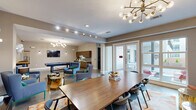 Located at 1010 S Pearl Expy in Dallas, TX, this single-floor community amenity space offers a thoughtfully designed, open-concept layout that blends entertainment and comfort seamlessly. The polished concrete floors and neutral warm-toned walls create a clean, modern backdrop throughout the entire space.

At the heart of the room sits a full-size billiards table beneath a striking mid-century modern chandelier with brass and globe accents. A wall-mounted flat-screen TV and wall sconces complete the game area, which is further supported by a wet bar and cabinetry along one wall — ideal for entertaining.

The lounge area features contemporary upholstered chairs with green-accented metal legs, arranged around sculptural gold side tables. Decorative circular wall art adds an artistic focal point, while track lighting provides flexible illumination across the space. A large dining table with seating for multiple guests anchors a defined gathering zone.

Floor-to-ceiling windows and French doors flood the interior with natural light and provide direct views and access to an outdoor courtyard patio, where additional seating and greenery can be seen. A connecting corridor leads to additional utility areas, maintaining a functional yet stylish flow throughout this well-appointed communal space.