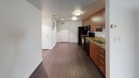 This single-floor unit offers a thoughtfully designed layout that balances function and style throughout its interconnected living spaces. Dark hardwood flooring runs consistently across the main areas, providing a sleek, cohesive aesthetic that ties the rooms together.

The kitchen is a standout feature, outfitted with rich warm-toned wood cabinetry extending floor to ceiling, offering generous storage. Granite countertops with gold and green tones provide ample prep space, complemented by a full suite of black appliances including an electric range with overhead microwave, dishwasher, and refrigerator. A stainless steel sink is positioned along the countertop run, and recessed and flush-mount lighting keep the space well-illuminated.

The open living area is characterized by a textured ceiling, bright white walls, and a distinctive exposed concrete structural column that adds an industrial architectural accent. A window allows natural light to filter into the space, enhancing the open feel. An adjacent walk-in closet provides practical storage off the main living zone.

A hallway connects the living and kitchen areas to the rest of the unit, leading to the bathroom. The bathroom features neutral gray walls, small-format tile flooring, a full-size soaking tub with shower, and a vanity, offering a clean and functional design.