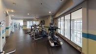 Located at 9240 Bruckhaus St in Raleigh, NC, this well-equipped fitness center offers a comprehensive workout environment across a single level. The space is thoughtfully designed with a distinctive color scheme featuring teal and soft yellow horizontal accent stripes along the walls, complemented by neutral tones and recessed lighting throughout.

The cardio zone is lined with large floor-to-ceiling windows that flood the area with natural light and provide views of the surrounding exterior, creating an open and energizing atmosphere. Elliptical machines, treadmills, and stair climbers are arranged to take full advantage of this bright setting.

The free weights area features a full rack of dumbbells, stability balls, and a weight bench, all reflected by a large wall-to-wall mirror that enhances the sense of space. An adjacent strength training section provides plate-loaded machines and additional benches, with wall-mounted televisions for entertainment during workouts.

A dedicated open floor area supports stretching and functional training exercises. Practical amenities include a water cooler station, hand sanitizer dispenser, and paper towel holder. The durable dark rubber flooring throughout is both functional and easy to maintain. The overall ambiance is clean, modern, and well-maintained, making this fitness center a fully functional facility for a wide range of workout routines.
