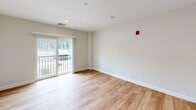 Located at 15 Factory St in Goffstown, NH, Unit 108 is a well-appointed single-floor unit that maximizes its compact footprint with smart design and modern finishes throughout. Light wood-tone flooring runs consistently across the living and kitchen areas, creating a cohesive and inviting atmosphere.

The kitchen is a standout feature, showcasing two-tone cabinetry with crisp white upper shaker-style cabinets and slate gray lower cabinets, complemented by bright white countertops and a full suite of stainless steel appliances, including a refrigerator, range, microwave, and dishwasher. A single-basin sink with a matte black faucet adds a contemporary touch.

The open living area is bright and airy, with neutral white walls and recessed lighting. A sliding glass door leads to a private balcony with outdoor views of the surrounding greenery, bringing in generous natural light. The entrance area features multiple closet spaces with classic two-panel white doors, offering substantial built-in storage.

The bathroom includes a full bathtub and shower with a curved black rod, tile flooring, and clean, understated finishes. The unit's overall design is clean, modern, and move-in ready, making it a functional and comfortable living space in a residential setting.