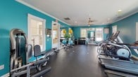 This fitness center is a comprehensively equipped workout facility designed to support a full range of exercise routines. The space is immediately distinguished by its bold teal walls, white crown molding, and crisp white window trim, creating an energetic yet clean aesthetic throughout. Durable dark rubber flooring covers the entire area, providing a practical and resilient surface ideal for all types of physical activity.

The cardio section features multiple treadmills with individual digital displays, positioned along a wall of large windows that frame views of the outdoor surroundings, including a pool area and surrounding greenery — offering an inviting backdrop during workouts. Ceiling fans are installed throughout, enhancing air circulation and comfort.

The strength training area is generously outfitted with a multi-station cable machine, a variety of selectorized weight machines targeting different muscle groups, and a well-stocked dumbbell rack. Exercise accessories such as stability balls in multiple sizes and adjustable benches round out the free-weight section.

Full-length mirrors are strategically placed to allow users to monitor their form. The space benefits from recessed lighting supplemented by abundant natural light streaming through multiple windows on opposing walls. A dedicated exit door provides convenient access to the outdoor area, seamlessly connecting the interior fitness space with the exterior amenities.