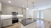 Located in Lewisville, TX, this thoughtfully designed one-bedroom, one-bathroom residence offers a cohesive modern aesthetic with high-quality finishes throughout its efficiently laid-out single floor. The open-concept kitchen serves as the centerpiece of the living area, showcasing a large island topped with dark granite countertops, an undermount sink with a brushed gold faucet, and pendant lighting with glass fixtures. Soft gray shaker-style cabinetry lines the walls, complemented by a textured tile backsplash and a full suite of stainless steel appliances, including a French door refrigerator, gas range, over-the-range microwave, and dishwasher. Gold hardware accents tie the kitchen's design together seamlessly. Light wood-look flooring runs through the main living areas, creating a warm, airy atmosphere. The bedroom offers comfortable carpeted flooring, a ceiling fan, and a window with blinds that allows natural light to filter in. A dedicated closet provides organized storage. The bathroom continues the home's refined design language with a gray vanity topped with granite, gold-framed mirror, gold fixtures, and wood-look tile flooring. Recessed lighting and a wall-mounted cabinet add both style and practicality to the space.