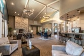 This expansive single-floor community space is defined by its dramatic vaulted ceilings and open, flowing layout that seamlessly connects multiple functional zones. The central gathering area features warm wood-look flooring, large decorative columns, and a statement chandelier that draws the eye upward toward the soaring ceiling. A round dining table with navy blue upholstered chairs anchors the dining area beneath an elegant hoop-style pendant light.

The fully equipped kitchen offers abundant white shaker cabinetry extending to the ceiling, granite countertops, a mosaic tile backsplash, stainless steel appliances including a refrigerator and dishwasher, and a breakfast bar with barstool seating — ideal for casual gatherings or catered events.

The lounge area is highlighted by a floor-to-ceiling stacked stone fireplace that serves as a commanding focal point, surrounded by comfortable seating and accented by deep teal accent walls that lend a bold, contemporary feel. Large windows throughout the space invite natural light and provide views of the surrounding outdoor areas, including a glimpse of a pool. A separate seating area with plush neutral sofas and armchairs offers a relaxed retreat. The overall ambiance is sophisticated yet welcoming, blending modern design elements with warm, residential-style comfort.