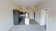 This single-floor space showcases a thoughtfully designed open-concept layout that seamlessly connects the kitchen and living areas. The kitchen is outfitted with dark charcoal cabinetry, stainless steel appliances including a refrigerator, microwave, and range, and is anchored by a sleek white central island with a built-in sink, ideal for both meal preparation and casual dining. Pendant lighting above the island adds a modern touch, complementing the flush-mount ceiling fixtures throughout. The flooring transitions from durable wood-look planks in the kitchen and entryway to plush carpet in the living area, providing both practicality and comfort. The neutral wall tones and clean lines throughout contribute to a bright, contemporary ambiance. Near the entryway, multiple doors suggest convenient access to closets or additional storage, adding to the functional design of the space. The overall aesthetic is clean and modern, combining dark and light contrasting elements for a polished, move-in-ready feel.
