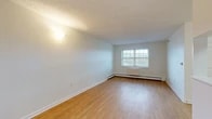 Located at 64 N De Baun Ave in Airmont, NY, this one-bedroom, one-bathroom residence offers a thoughtfully organized single-floor layout with a clean, neutral aesthetic throughout. The open living and dining area is bright and welcoming, featuring warm hardwood-style flooring, a textured ceiling, and a large double-hung window that invites natural light and outdoor views. A wall-mounted light fixture adds a soft glow to the space, while baseboard heating runs along the perimeter for efficient climate control. The kitchen is positioned adjacent to the living area with a pass-through counter and breakfast bar, wood-tone cabinetry, and white appliances, creating a functional and connected space ideal for everyday living. The bedroom is comfortably sized, finished with plush carpeting, a window with blinds, and direct access to a closet — with a total of three closets spread throughout the unit providing generous storage options. The bathroom features a full tub and shower combination equipped with stainless steel grab bars, white tile surround, and a wood-tone vanity. The overall ambiance is practical and understated, with a neutral color palette that offers a move-in-ready canvas in the charming hamlet of Airmont, New York.