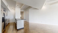 This single-floor space showcases a contemporary industrial-modern design throughout. Polished concrete floors extend seamlessly across all areas, lending a cohesive, urban aesthetic that pairs with crisp white walls and exposed structural beams on the ceiling.

The open-concept kitchen and living room serve as the heart of the space. The kitchen is outfitted with sleek gray cabinetry, dark granite countertops, a mosaic tile backsplash, stainless steel appliances including a built-in microwave and range, and a white refrigerator with ice dispenser. A central island with matching granite provides additional prep space and casual seating. The living area flows openly from the kitchen, benefiting from large floor-to-ceiling windows that invite abundant natural light and views of the surrounding cityscape.

The bathroom features a deep soaking tub surrounded by striking black mosaic tile, offering a bold contrast to the otherwise neutral palette. A hallway leads to the additional rooms, including a generously sized walk-in closet outfitted with wire shelving on three walls for ample storage. The unclassified room presents a versatile, well-lit space suitable for use as a bedroom or office. Throughout the space, recessed and track lighting complements the natural light, creating a bright, airy atmosphere.