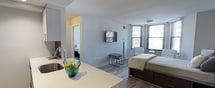 This single-floor studio is designed with an open-concept layout that seamlessly integrates the kitchen, living, and sleeping areas into one cohesive, functional space. The modern kitchen features sleek white cabinetry, a stainless steel sink set into a clean quartz countertop peninsula, and built-in appliances — providing ample prep space and storage without overwhelming the room. The peninsula serves as a natural dividing element between the kitchen and the rest of the living space, creating distinct zones while maintaining an open and airy feel. Light gray walls and light-toned wood-style flooring run throughout, lending a consistent, contemporary aesthetic. The sleeping area accommodates a full-sized bed and is complemented by a wall-mounted flat-screen television, making the space both restful and functional. A second sleeping or sitting alcove is visible through an open doorway, offering additional flexibility. Large windows fitted with horizontal blinds allow generous natural light to fill the interior, enhancing the sense of openness. A desk and side table provide a dedicated workspace, rounding out the practical amenities. Overall, the space offers a clean, modern, and efficiently organized environment suited for comfortable everyday living.