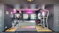 Located at 9001 SW 122nd Ave in Miami, Florida, this single-floor fitness facility offers a comprehensive workout experience combining a well-appointed gym with a dedicated racquetball court. The facility is accessed through glass double doors that invite natural light into the entry area, which features pendant lighting, a seating bench, and a rubberized floor surface ideal for high-traffic use.

The main workout area is boldly designed with dark charcoal and deep purple walls, accented by a vibrant pink neon "You Can Do It" sign that sets a motivating tone. Large windows provide natural light and views of the surrounding greenery. The gym is thoroughly equipped with treadmills, elliptical machines, stationary and spin bikes, a rowing machine, a multi-station cable machine, a full dumbbell rack, medicine balls, and additional strength training equipment. Full-length mirrors line the walls to support proper form and enhance the sense of space. Flooring transitions between rubberized mat sections and smooth hardwood, suited to various workout activities.

Adjacent to the gym, the racquetball court features a classic hardwood floor with regulation red boundary lines, bright white walls, and soaring ceilings, offering a clean and professional playing environment. The court is accessible directly from the entry vestibule via a glass-paneled door, seamlessly connecting both fitness spaces.