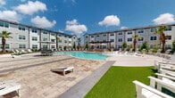 This contemporary multi-story residential community is designed around an expansive, resort-style outdoor amenity space that serves as its focal point. The property's architecture features a clean, modern aesthetic with grey horizontal siding, large multi-pane windows, and pitched rooflines that lend a polished, well-maintained appearance across all floors.

At the heart of the community lies a generously sized freeform swimming pool with crystal-clear blue water, surrounded by a broad, paver-tiled sundeck. The deck is well-appointed with white lounge chairs, patio umbrellas, and shaded seating areas, providing ample space for relaxation and socializing. A pergola structure adds an architectural accent to the outdoor entertaining zone, while a separate leisure lawn area features artificial turf and cornhole boards for recreational activities.

The landscaping throughout the courtyard is thoughtfully curated, with tall palm trees, flowering plants, ornamental grasses, and layered shrubbery that create a vibrant, tropical ambiance. Wrought-iron fencing with gated walkways connects the buildings to the pool area, ensuring both accessibility and security. A clubhouse-style structure with arched entryways, stone detailing, and large glass doors borders the pool, providing an indoor-outdoor connection. The overall environment is bright, open, and inviting, with abundant natural light and open sky views enhancing the outdoor living experience.
