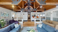 Located at 5680 Hibernia Dr in Columbus, OH, this remarkable single-floor living space makes an immediate impression with its dramatic vaulted ceiling supported by exposed heavy-timber trusses with steel gusset plate connectors — a bold architectural statement that defines the entire interior. Warm wood-paneled ceilings and rich hardwood flooring run throughout, creating a cohesive and inviting atmosphere.

The living room serves as the heart of the space, centered around a floor-to-ceiling stacked stone fireplace flanked by large windows that frame lush, tree-lined outdoor views. The open layout flows seamlessly between a casual seating area, a sunroom-style windowed nook bathed in natural light, and a stepped transition toward a kitchen and dining area visible in the background. The kitchen features bar-height seating with vibrant orange stools, white cabinetry, and stainless steel appliances.

A ceiling fan complements the soaring peak overhead, while track lighting illuminates the space throughout. Interior railings with white balustrades subtly define level changes, adding architectural interest. Every corner of this space balances rustic character with clean, modern touches, resulting in an open, airy, and uniquely designed interior full of warmth and personality.