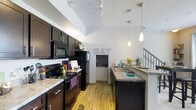 This two-story residence offers a thoughtfully designed layout that combines modern aesthetics with functional living. The kitchen is a standout feature, boasting rich dark-stained cabinetry that contrasts beautifully with light stone countertops and warm hardwood-style flooring. Vaulted ceilings soar overhead, creating a dramatic sense of height and openness that fills the space with an airy, expansive feel. Black appliances, including a refrigerator, range, and built-in microwave, lend a sleek, contemporary look to the cooking area. Elegant pendant lighting hangs above the countertop, adding both task illumination and a stylish finishing touch. A dedicated sink area with additional counter space runs along one side, providing ample room for meal preparation. The kitchen flows naturally into the adjoining dining area, subtly marked with an "EAT" wall accent, reinforcing the space's casual yet inviting character. Track lighting along the ceiling further enhances the brightness of the room. A staircase, visible at the far end of the kitchen, leads to the second floor, integrating the two levels seamlessly. Throughout the space, the combination of high ceilings, quality finishes, and an open layout creates a welcoming and functional home environment.