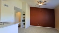 This single-story space presents a thoughtfully designed interior that balances architectural detail with a warm color palette. The main living area is anchored by a striking terracotta-red accent wall, which adds depth and visual interest against the surrounding cream-toned walls. A ceiling fan with a light fixture is centrally mounted overhead, providing both comfort and ambient lighting to the room.

The flooring throughout is covered in neutral-toned carpet, contributing to the space's cozy and comfortable feel. A key architectural highlight is the built-in arched niche unit, featuring multiple shelving tiers framed by a classic arch detail — ideal for displaying decorative items or plants, as shown by the greenery and décor currently adorning the shelves.

Adjacent to the living area, a partial wall with a countertop surface suggests a pass-through or breakfast bar, likely connecting to a kitchen space beyond. The arched doorway visible in the background reinforces the consistent use of curved architectural elements throughout the design, lending the space a cohesive and elegant character. Overall, the space offers a warm, inviting ambiance with distinctive design touches.