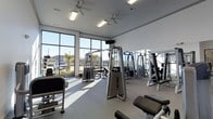This single-story fitness center is designed to provide a comfortable and functional workout environment. The space is defined by expansive floor-to-ceiling windows along the exterior wall, flooding the interior with abundant natural light and offering unobstructed views of the surrounding landscape, including palm trees and the neighboring streetscape. The bright white walls and light-toned flooring further enhance the open, airy atmosphere.

The facility is outfitted with a comprehensive selection of professional-grade Precor strength training machines, strategically arranged to allow for comfortable circulation between equipment. A dedicated dumbbell rack is visible, complementing the variety of resistance machines available. Large mirrors are positioned along one wall, a practical feature that supports proper form during workouts while also visually expanding the space.

Overhead, multiple ceiling fans are evenly distributed throughout the room, working in conjunction with the fluorescent lighting to maintain a well-lit and well-ventilated environment. The combination of ample natural light, quality equipment, and thoughtful layout makes this fitness center a practical and inviting space for a wide range of exercise routines.