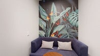 Located at 701 Richmond Ave in Houston's vibrant Montrose district, this intimate lounge space is a single, thoughtfully curated room that makes a strong visual impact despite its compact footprint. The design aesthetic balances bold artistic expression with clean, modern simplicity.

The defining feature of the room is a striking floor-to-ceiling tropical wallpaper mural depicting lush bird-of-paradise flowers, monstera leaves, and banana foliage rendered in vivid teals, oranges, and reds against a deep, dark background. This dramatic botanical backdrop anchors the seating area, where a modern blue sofa with striped accent pillows is paired with a warm-toned circular wood coffee table.

The flooring features a deep blue-purple marbled tile pattern that complements the room's cool color palette and adds a sense of depth and texture underfoot. The remaining walls are finished in crisp white, keeping the space feeling open and uncluttered. A framed black-and-white fine art photograph adorns one wall near the entrance, contributing to the gallery-like quality of the space.

Access is provided through a dark-framed door with a frosted glass panel, offering both privacy and a refined architectural detail. Overall, the space presents a cohesive, design-forward environment ideal for relaxed, private gatherings or a quiet retreat.