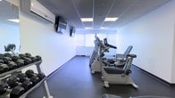 This single-floor fitness room is designed to offer a functional and focused exercise environment. The space is outfitted with a selection of cardio equipment, including stationary bikes, an elliptical trainer, and a treadmill, catering to a variety of workout preferences. The flooring is finished in durable, dark rubber matting — a practical choice that provides cushioning and stability during exercise. Overhead, a drop ceiling with recessed panel lighting ensures consistent, even illumination throughout the room. A wall-mounted split air conditioning unit provides dedicated climate control, keeping the environment comfortable regardless of the intensity of activity. Two wall-mounted flat-screen televisions are positioned for easy viewing from the cardio machines, enhancing the overall workout experience. Natural light filters into the space through a set of windows dressed with vertical blinds, offering a pleasant connection to the outdoors while maintaining privacy. The walls are finished in clean white, contributing to a bright and open atmosphere. The overall design is straightforward and practical, prioritizing utility and ease of use for fitness activities.