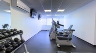 This single-floor fitness room is designed to offer a functional and focused exercise environment. The space is outfitted with a selection of cardio equipment, including stationary bikes, an elliptical trainer, and a treadmill, catering to a variety of workout preferences. The flooring is finished in durable, dark rubber matting — a practical choice that provides cushioning and stability during exercise. Overhead, a drop ceiling with recessed panel lighting ensures consistent, even illumination throughout the room. A wall-mounted split air conditioning unit provides dedicated climate control, keeping the environment comfortable regardless of the intensity of activity. Two wall-mounted flat-screen televisions are positioned for easy viewing from the cardio machines, enhancing the overall workout experience. Natural light filters into the space through a set of windows dressed with vertical blinds, offering a pleasant connection to the outdoors while maintaining privacy. The walls are finished in clean white, contributing to a bright and open atmosphere. The overall design is straightforward and practical, prioritizing utility and ease of use for fitness activities.