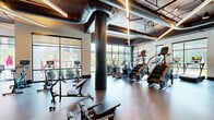This expansive fitness center offers a well-appointed, single-floor layout designed to accommodate a full range of workout activities. The space is defined by its industrial-chic aesthetic, featuring exposed ductwork and structural columns, high ceilings with linear accent lighting, and durable speckled rubber flooring throughout. Large floor-to-ceiling windows line the perimeter, flooding the interior with natural light and providing open views of the surrounding greenery and streetscape.

The facility is organized into distinct functional zones. A dedicated cardio area houses a comprehensive selection of equipment, including StairMaster climbers, ellipticals, and stationary cycles, all positioned to take full advantage of the expansive window views. A separate strength training zone offers a robust selection of cable machines, weight racks, and free weights, complemented by mirrored walls that enhance the sense of space. An additional enclosed studio area, accessible through sleek steel-framed glass doors, provides a versatile space for group classes or private training sessions.

The entry area features a polished aggregate floor and clean architectural details, setting an professional tone for the facility. Throughout, the combination of contemporary finishes, thoughtful equipment layout, and ample natural light creates an energizing and highly functional environment suited for serious fitness pursuits.