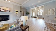 This refined single-floor communal space blends classic architectural details with a warm, contemporary aesthetic. Light hardwood flooring stretches throughout the open-concept layout, reflecting natural light that pours in through large arched and multi-pane windows dressed with bold, patterned drapery.

The main gathering area is anchored by a generous seating arrangement featuring a plush sofa accented with colorful throw pillows, complemented by wingback accent chairs and layered area rugs with geometric patterns. Tray ceilings with recessed lighting and elegant crown molding add a sense of refinement and volume to the space. Wall sconces with gold-toned fixtures provide warm ambient lighting throughout.

A standout architectural feature is the arched pass-through that frames a stylish bar counter finished in white wainscoting, topped with light stone and served by gold-accented bar stools. Open shelving behind the bar displays decorative accents, creating a sophisticated focal point.

Near the entrance, a welcoming seating nook with two wingback chairs, a geometric-patterned rug, and a round accent table sits beside oversized glass-panel entry doors, offering views to the outdoors. A pendant lantern in a brushed gold finish hangs above, adding a polished finishing touch. The overall ambiance is elegant yet inviting, ideal for communal gathering and relaxation.