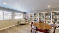 This charming community library in Escondido, California, offers a welcoming and functional space designed for relaxed reading and browsing. The single-floor layout is thoughtfully organized to maximize comfort and accessibility, featuring durable wood-look flooring complemented by textured carpet tiles that add warmth underfoot.

The main reading area is anchored by a set of full-height white built-in bookshelves stocked with a wide variety of titles, offering a rich selection for visitors. A cushioned accent chair and side table are positioned nearby, creating an inviting reading nook bathed in natural light from the windows. Additional seating includes upholstered lounge chairs and a central wooden table with dining-style chairs, providing flexible spaces for both individual and group use.

A built-in granite countertop along one wall serves as a convenient check-in or resource station, framed by an elegant arched opening with glass-panel windows that visually connect adjacent areas. French doors with glass panes allow light to flow through while maintaining a sense of openness. Recessed lighting and crown molding throughout lend a clean, polished finish to the space.

A wall-mounted HVAC unit ensures year-round comfort, while exterior windows provide views of the surrounding neighborhood. The overall ambiance is calm, organized, and community-oriented.