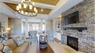 Located at 3400 Briercliff Road in Greenville, North Carolina, this single-story space offers a thoughtfully designed interior that balances warmth and sophistication. The main living area immediately draws attention with its coffered ceiling, accented by rich wood planking and illuminated by a statement chandelier with a wrought-iron and gold finish, adding a refined yet inviting ambiance.

A floor-to-ceiling stacked stone fireplace serves as the room's focal point, flanked by built-in cabinetry and shelving in crisp white, providing both functional storage and visual balance. Hardwood flooring runs throughout, complementing the neutral gray walls and creating a cohesive, timeless aesthetic.

The seating arrangement, featuring soft blue accent chairs and a neutral sofa, encourages a relaxed and comfortable atmosphere. Decorative columns subtly define the transition between the main living space and an adjoining area, which benefits from abundant natural light streaming through large windows and glass-paneled entry doors. Recessed lighting throughout ensures the space remains bright and welcoming at all hours, making it an ideal environment for gathering and socializing.