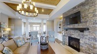 Located at 3400 Briercliff Road in Greenville, North Carolina, this single-story space offers a thoughtfully designed interior that balances warmth and sophistication. The main living area immediately draws attention with its coffered ceiling, accented by rich wood planking and illuminated by a statement chandelier with a wrought-iron and gold finish, adding a refined yet inviting ambiance.

A floor-to-ceiling stacked stone fireplace serves as the room's focal point, flanked by built-in cabinetry and shelving in crisp white, providing both functional storage and visual balance. Hardwood flooring runs throughout, complementing the neutral gray walls and creating a cohesive, timeless aesthetic.

The seating arrangement, featuring soft blue accent chairs and a neutral sofa, encourages a relaxed and comfortable atmosphere. Decorative columns subtly define the transition between the main living space and an adjoining area, which benefits from abundant natural light streaming through large windows and glass-paneled entry doors. Recessed lighting throughout ensures the space remains bright and welcoming at all hours, making it an ideal environment for gathering and socializing.