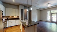 This single-floor residence offers a thoughtfully designed open-concept layout that seamlessly connects the kitchen, dining, and living areas. The kitchen is a standout feature, showcasing dark granite countertops, crisp white cabinetry, a subway tile backsplash, and a full suite of stainless steel appliances including a refrigerator, range, and microwave. A central island with a double sink and stylish pendant lighting serves as both a functional workspace and a visual focal point. Light hardwood flooring runs through the kitchen and living areas, while the living room impresses with a tray ceiling detailed with crown molding and French doors that open to an outdoor space, allowing for an abundance of natural light. The bedrooms are carpeted for comfort and each features windows with exterior views. The bathrooms are finished with granite-topped vanities, white cabinetry, large mirrors, tile flooring, and tub-shower combinations with white subway tile surrounds. Crown molding and consistent neutral tones throughout contribute to a cohesive, polished ambiance, making this a refined and functional single-level home.