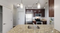 This two-story residence offers a thoughtfully designed layout spread across two levels, combining modern finishes with functional living spaces. The kitchen serves as a central highlight, featuring rich dark-wood cabinetry, light granite countertops, a stainless steel undermount sink set into an island or peninsula, and a full suite of stainless steel appliances including a refrigerator, electric range, and over-the-range microwave. Stylish pendant lighting and a track lighting system illuminate the space with a contemporary flair. The main living area showcases warm hardwood flooring and large windows fitted with horizontal blinds, allowing an abundance of natural light to fill the room. A carpeted staircase with a warm wood handrail and crisp white baseboards connects the two levels. Upstairs, the bedrooms are finished with plush neutral carpeting, large windows, and generous closet space equipped with wire shelving and hanging rods. The bathrooms are finished with large-format tile surrounding the tub-shower combinations, granite-topped dark-wood vanities, and tile flooring, creating a cohesive and polished aesthetic throughout. The overall design balances clean, neutral tones with warm wood accents for a comfortable and modern ambiance.