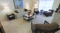 This single-floor space centers around a thoughtfully designed living room that balances comfort and classic elegance. The sitting area is anchored by a generously sized skirted sofa in a soft neutral tone, complemented by a pair of dark-upholstered armchairs that add contrast and depth to the overall palette. A sleek glass-topped coffee table with a dark metal frame serves as the focal point of the seating arrangement, providing both functionality and a refined aesthetic. Warm ambient lighting is provided by traditional table lamps positioned throughout the room, casting a soft, inviting glow across the space. A large-scale impressionist-style painting adorns one wall, adding a vibrant splash of color and artistic character. Natural light filters in through a window, contributing to the room's bright and airy feel. The neutral carpeted flooring ties the space together cohesively, while carefully placed side tables and decorative accessories — including books, vases, and sculptural accents — enhance the room's curated, welcoming atmosphere. The overall ambiance is warm, timeless, and well-balanced.