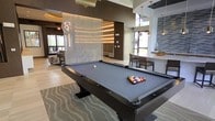 This contemporary amenity lounge is thoughtfully designed to offer a dynamic social environment with a variety of gathering spaces. At the heart of the room sits a full-size billiards table with a sleek dark finish, set atop a patterned area rug that anchors the space with a touch of texture and style. Overhead, an elegant globe pendant light adds a sculptural element to the room's refined aesthetic.

A striking decorative curtain wall of shimmering strands serves as a dramatic focal point, dividing the space while maintaining an open, airy feel. The high ceiling further enhances the sense of volume, while large windows allow natural light to pour in, brightening the interior and offering views to the outdoors.

Adjacent to the billiards area, a dining and bar-height seating section is outfitted with modern chairs and a light-toned countertop, illuminated by warm gold pendant fixtures. The design palette blends rich brown and neutral tones with reflective surfaces and layered lighting to create an upscale, welcoming atmosphere.

A comfortable lounge seating area with plush furnishings rounds out the space, making it ideal for relaxed socializing. Throughout, the finishes are polished and cohesive, reflecting a high standard of design suited for communal entertainment and leisure.