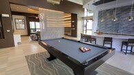 This contemporary amenity lounge is thoughtfully designed to offer a dynamic social environment with a variety of gathering spaces. At the heart of the room sits a full-size billiards table with a sleek dark finish, set atop a patterned area rug that anchors the space with a touch of texture and style. Overhead, an elegant globe pendant light adds a sculptural element to the room's refined aesthetic.

A striking decorative curtain wall of shimmering strands serves as a dramatic focal point, dividing the space while maintaining an open, airy feel. The high ceiling further enhances the sense of volume, while large windows allow natural light to pour in, brightening the interior and offering views to the outdoors.

Adjacent to the billiards area, a dining and bar-height seating section is outfitted with modern chairs and a light-toned countertop, illuminated by warm gold pendant fixtures. The design palette blends rich brown and neutral tones with reflective surfaces and layered lighting to create an upscale, welcoming atmosphere.

A comfortable lounge seating area with plush furnishings rounds out the space, making it ideal for relaxed socializing. Throughout, the finishes are polished and cohesive, reflecting a high standard of design suited for communal entertainment and leisure.